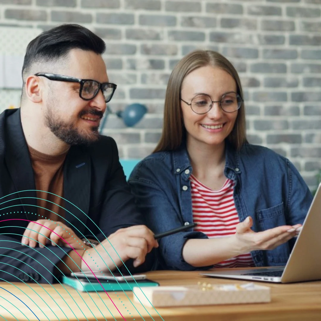 A man wearing glasses and a blazer is sitting beside a woman in glasses and stripy top. They are smiling and looking at a laptop.